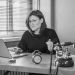 Black and white photo of a woman working at a desk with a laptop and vintage telephone.