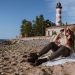 A couple enjoys a beach day by a lighthouse, with binoculars and a book for recreation.